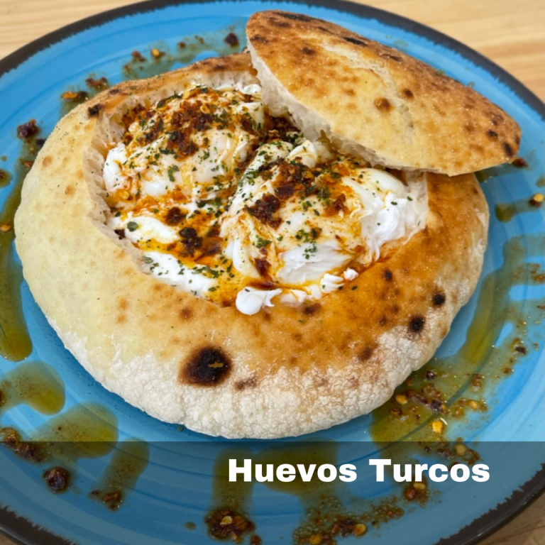 Dos huevos escalfados sobre una base de yogur cremosa, aderezado con salsa de mantequilla al pimentón, copos de chile y hierbas frescas, acompañado por pan.